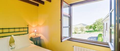 A bedroom with a bed, a nightstand with a lamp, a window with a view of the outside, and wooden beams on the ceiling.