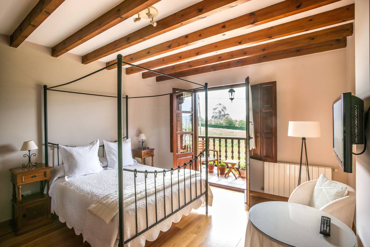Quarto com cama de dossel, teto de madeira, varanda com plantas em vasos e televisão na parede.