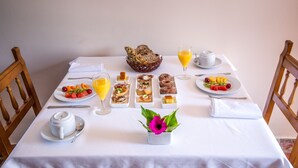 Una mesa de desayuno con diversos platos, un plato de fruta, un vaso de zumo de naranja y un pequeño arreglo floral.