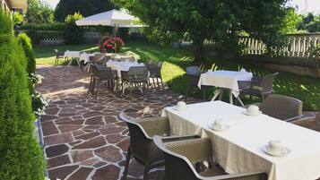 Une terrasse extérieure avec un sol en pierre, des tables recouvertes de nappes blanches, des chaises en osier et des parasols pour créer de l’ombre.