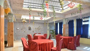 A room with a high ceiling, exposed beams, and a rustic wall design. There are tables and chairs covered with red tablecloths, and the room has large windows with blue curtains. - Hotel Siddarth Palace (Mangaldai)