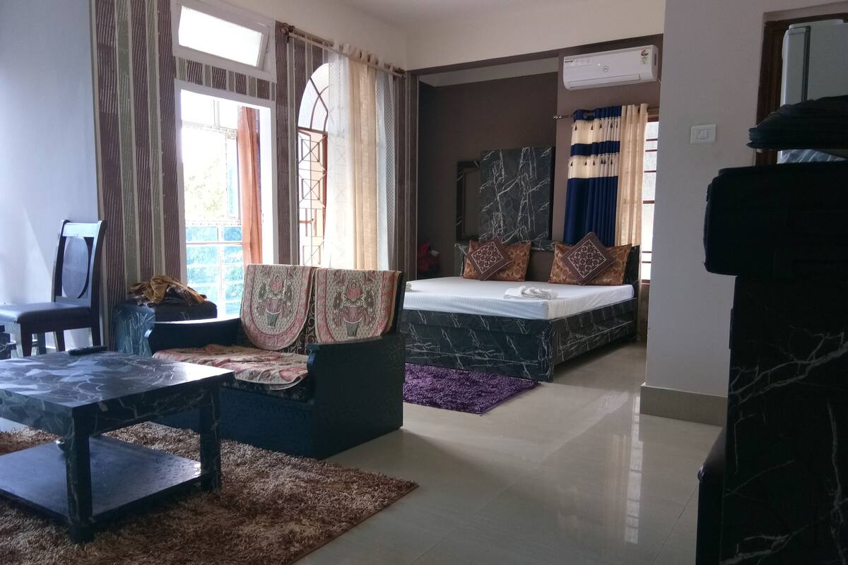 A living room with a sofa, a coffee table, a bed with patterned pillows, and a window with curtains.