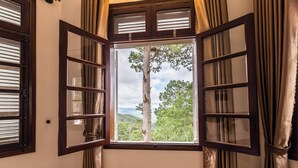 A room with a large window featuring wooden shutters, a view of a tree and mountains. - Dragon Dalat Villa (Xuan Huong - Da Lat)