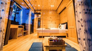A cozy wooden cabin room with a bed, a small table with snacks, and a window with a view of trees. - Dragon Dalat Villa (Xuan Huong - Da Lat)