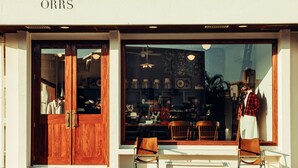 A storefront with a large window displaying various items, including a mannequin dressed in a plaid shirt and white skirt. - Orrs (Chatan)