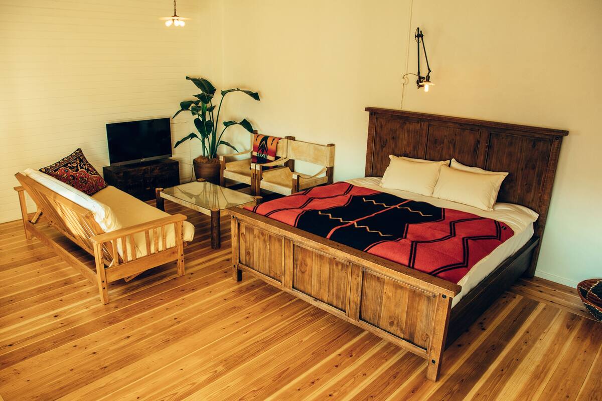 A wooden bed with a red and black blanket, a wooden chair, a television, and a potted plant in a room with wooden flooring.