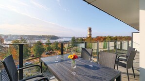 Een balkon met een tafel en stoelen, met uitzicht op een stadslandschap met een rivier en een toren.