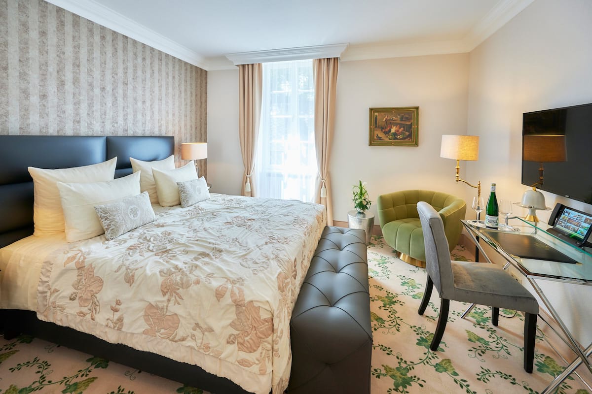 Una habitación de hotel con una cama grande, un sillón verde, un escritorio con una computadora y un televisor.