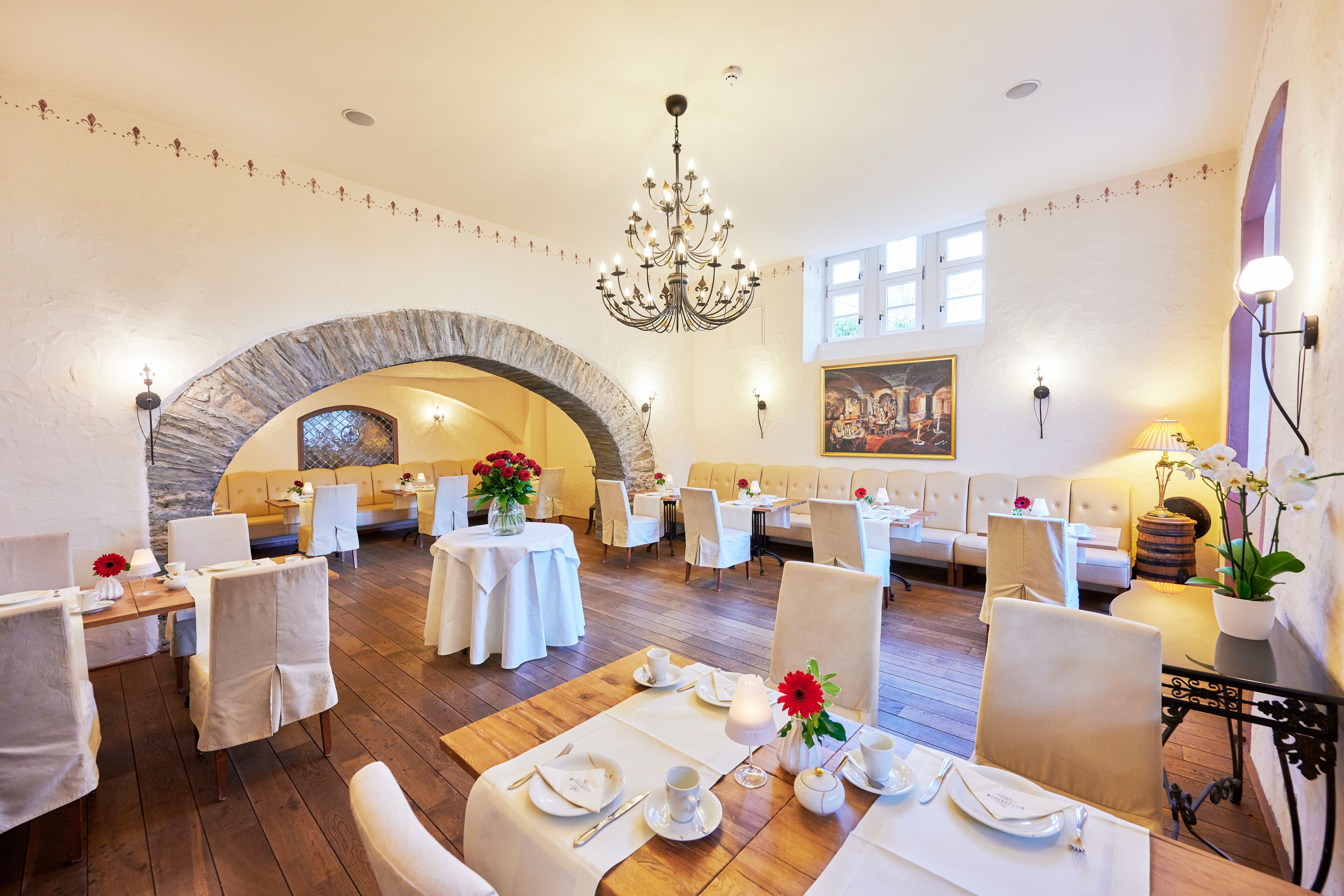 Un comedor con mesas dispuestas para el servicio, una araña y una estructura de piedra en arco al fondo.