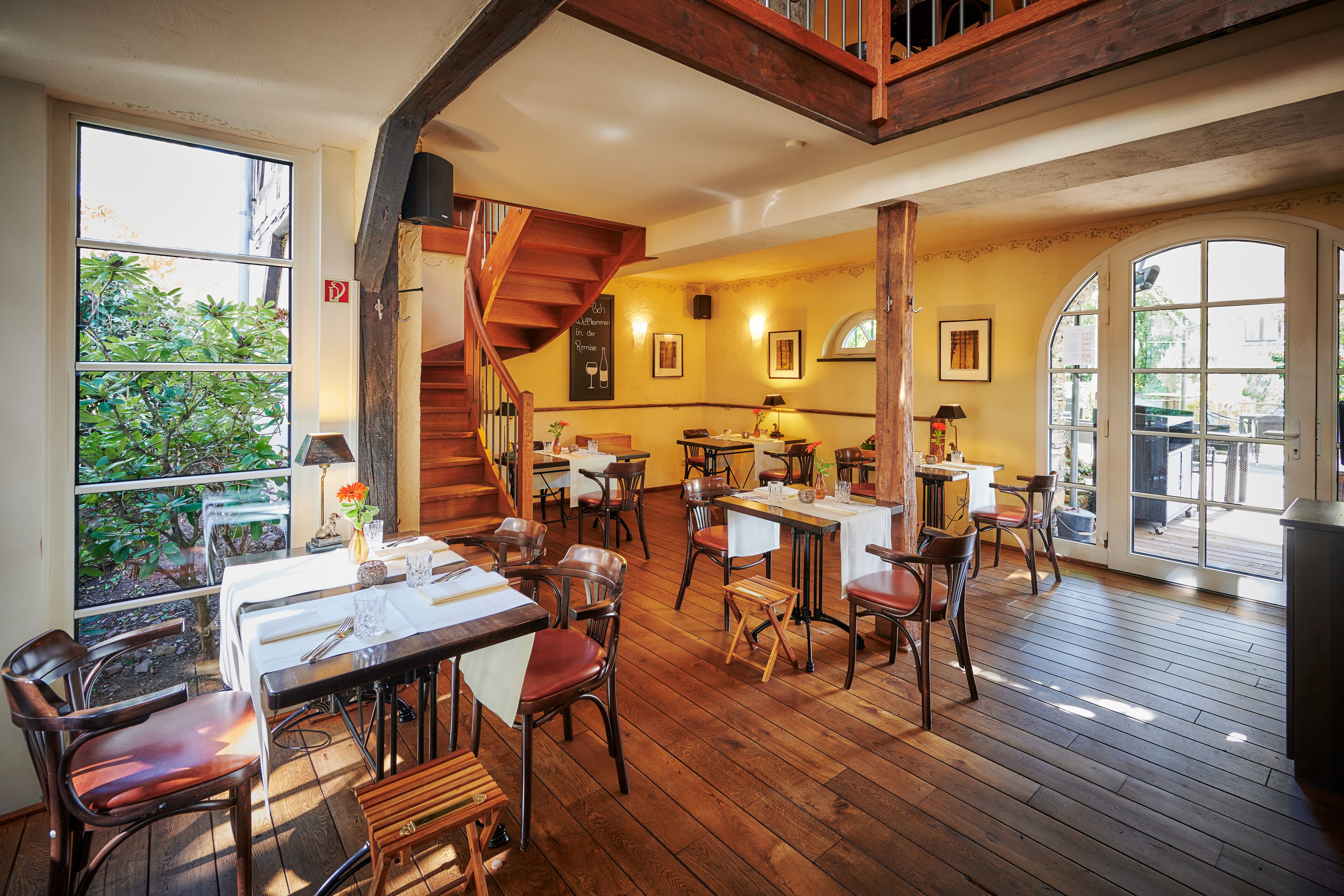 A restaurant with wooden floors, multiple tables and chairs, a staircase, and large windows offering a view of greenery.