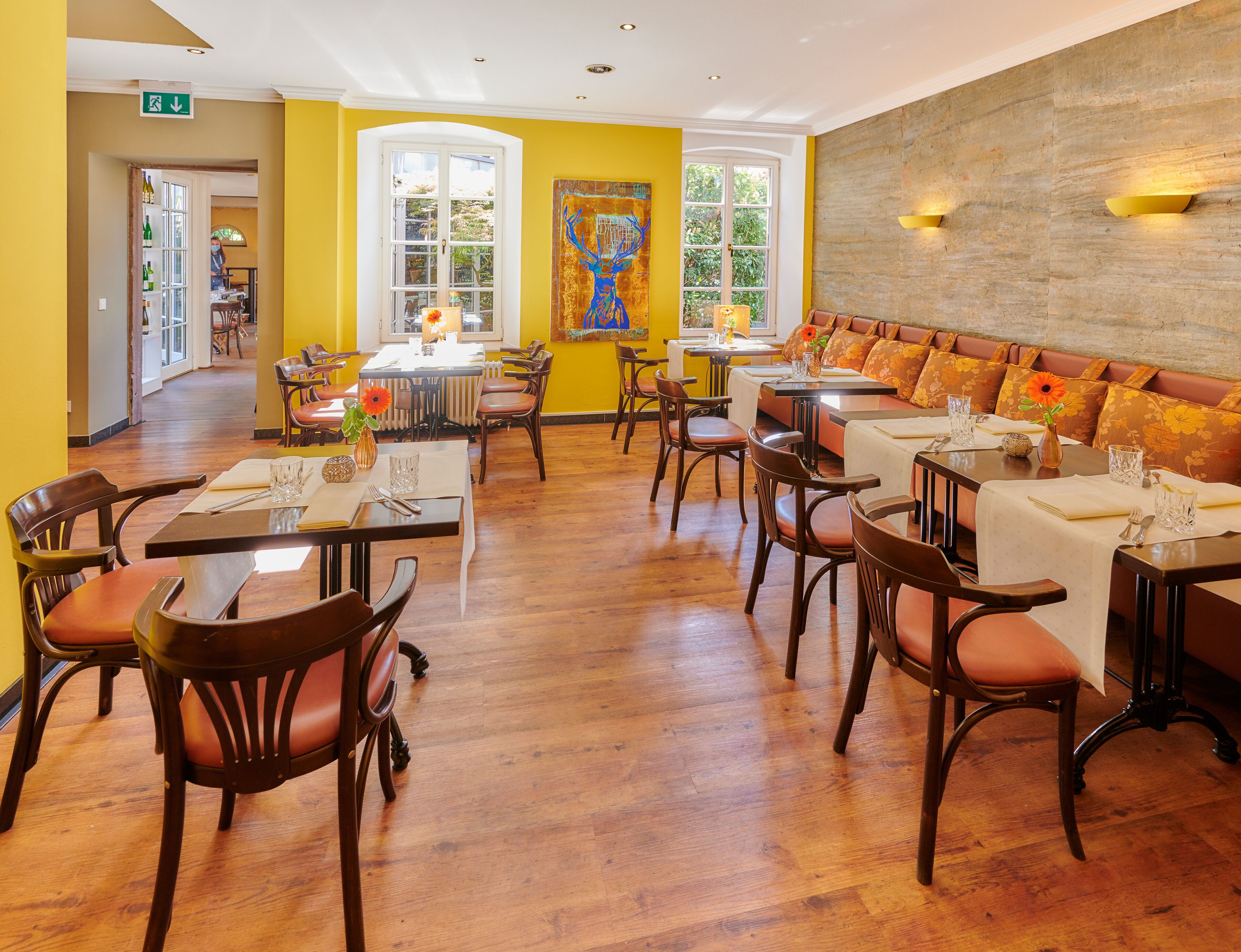 A restaurant with wooden flooring, yellow walls, and a large mural. Tables are set with white tablecloths and orange flowers.