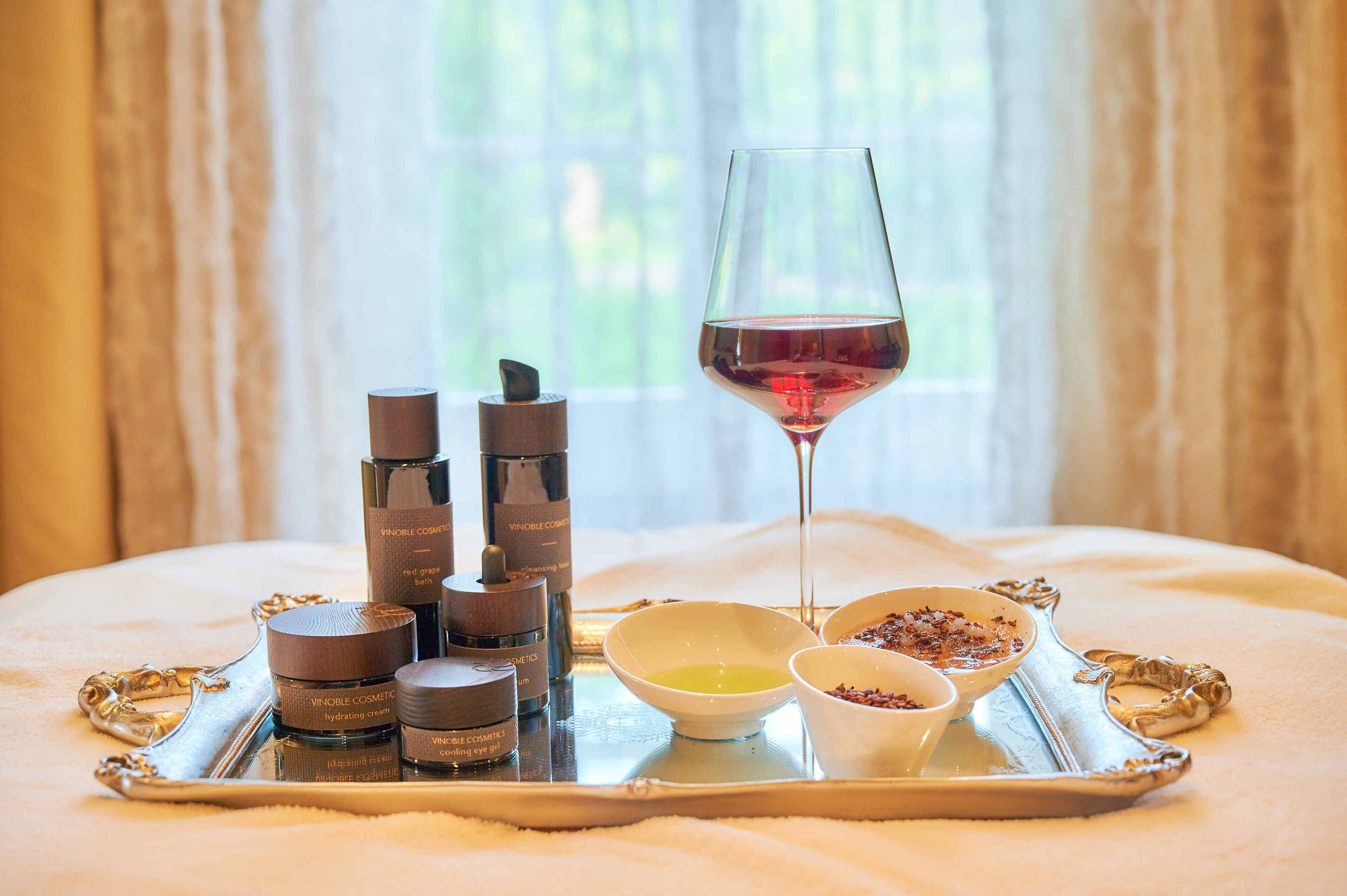 Uma bandeja com produtos de cuidados para a pele, uma taça de vinho e potes de creme sobre uma cama.