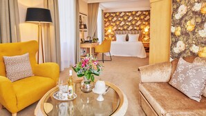 Une chambre d’hôtel avec un fauteuil jaune, une table basse ronde sur laquelle se trouvent une bouteille et des verres, un canapé et un lit dont le mur est recouvert d’un papier peint à motifs floraux.
