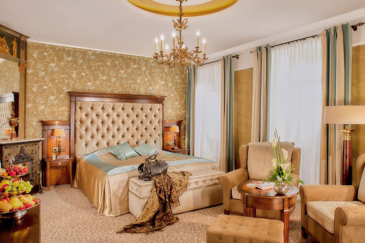 Un dormitorio lujoso con un cabecero capitonado, una cama grande, una zona de estar con sofá y sillón, una mesita de noche con lámpara y una cesta de frutas.