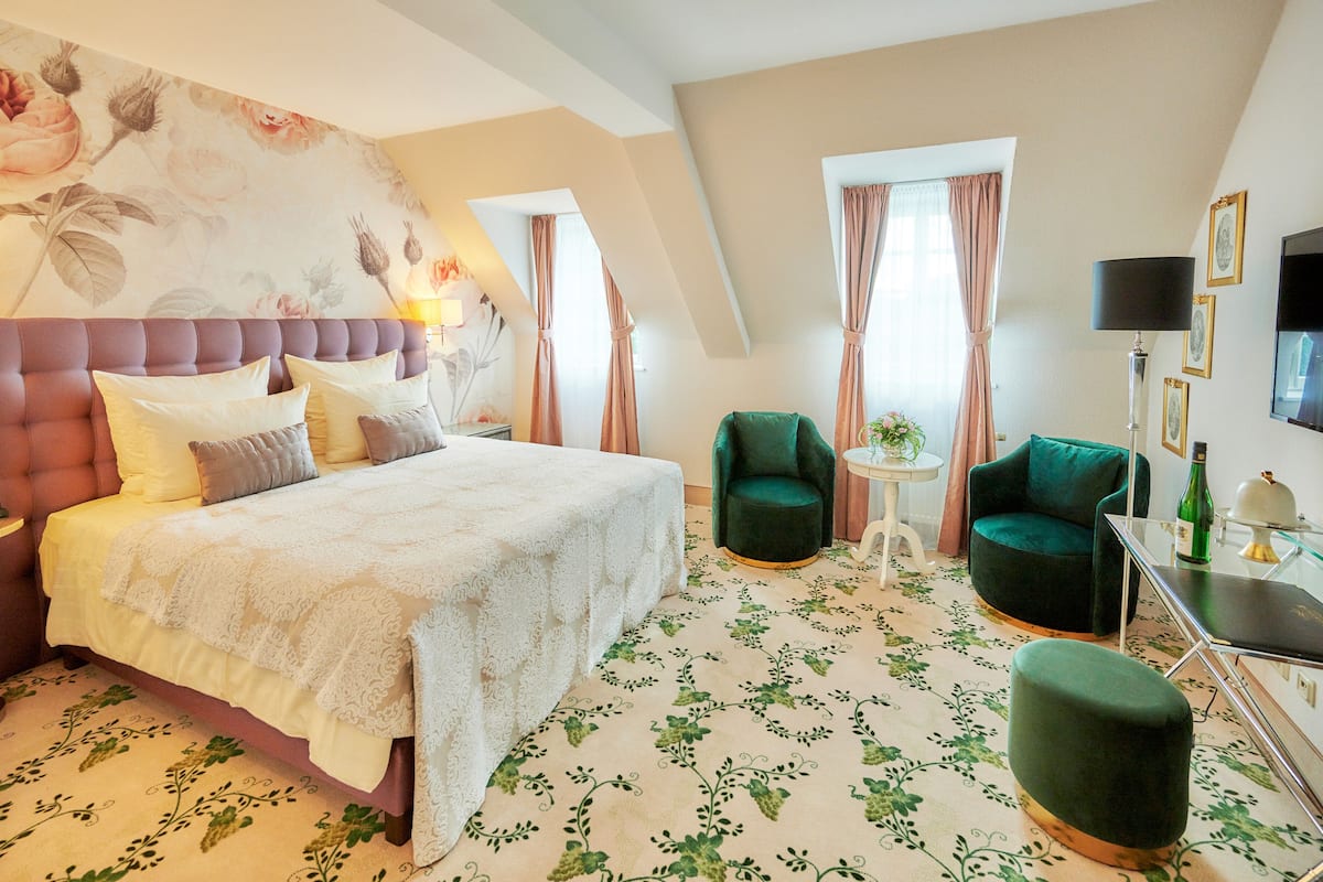 Un dormitorio con una cama grande, un sillón verde, una mesita y un televisor en la pared.