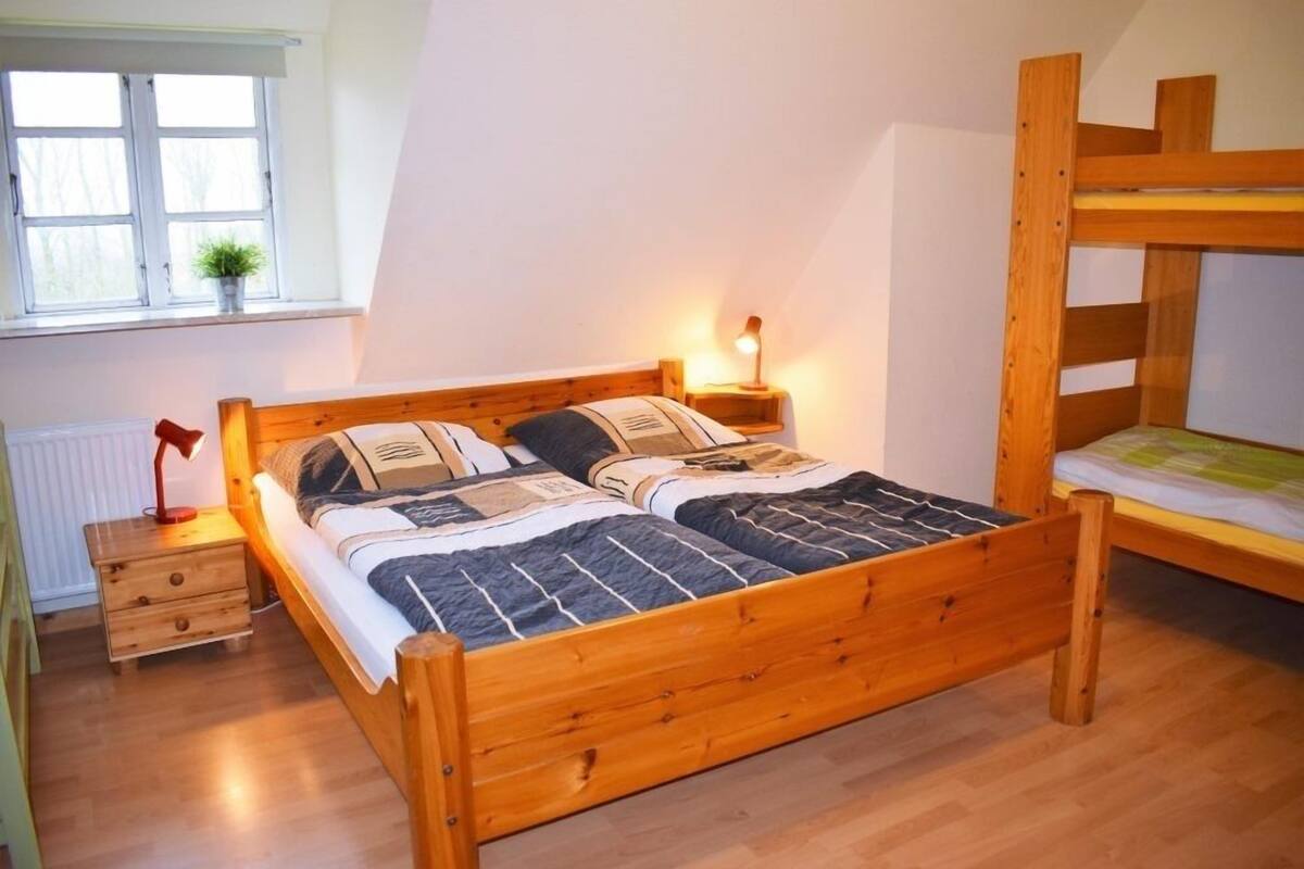 Un lit superposé en bois, avec une table de chevet et une lampe, une fenêtre avec une plante et un radiateur.
