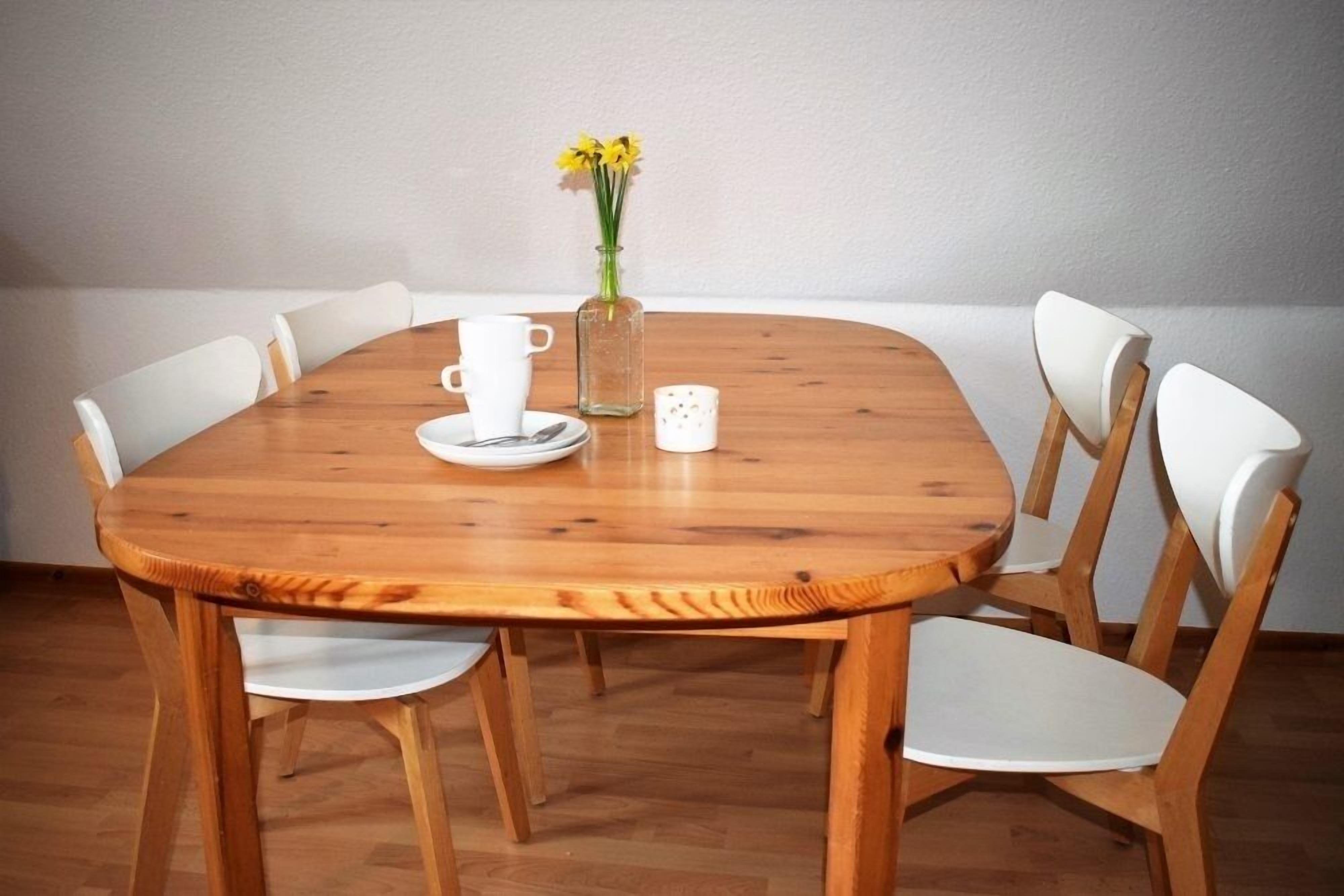 A wooden dining table with six white chairs, a vase with yellow flowers, and a white mug on the table.
