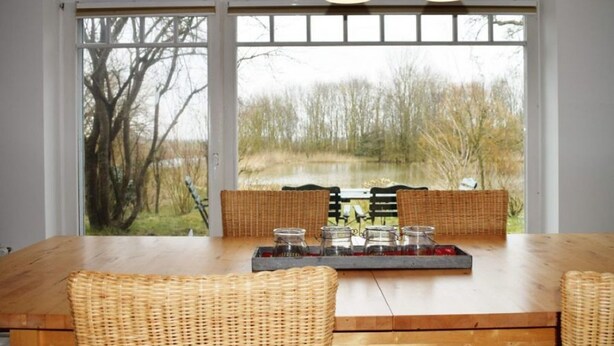 A dining area with a wooden table, wicker chairs, and a view of a natural landscape through large windows. - Reetdachhaus Bongsiel (Ockholm)