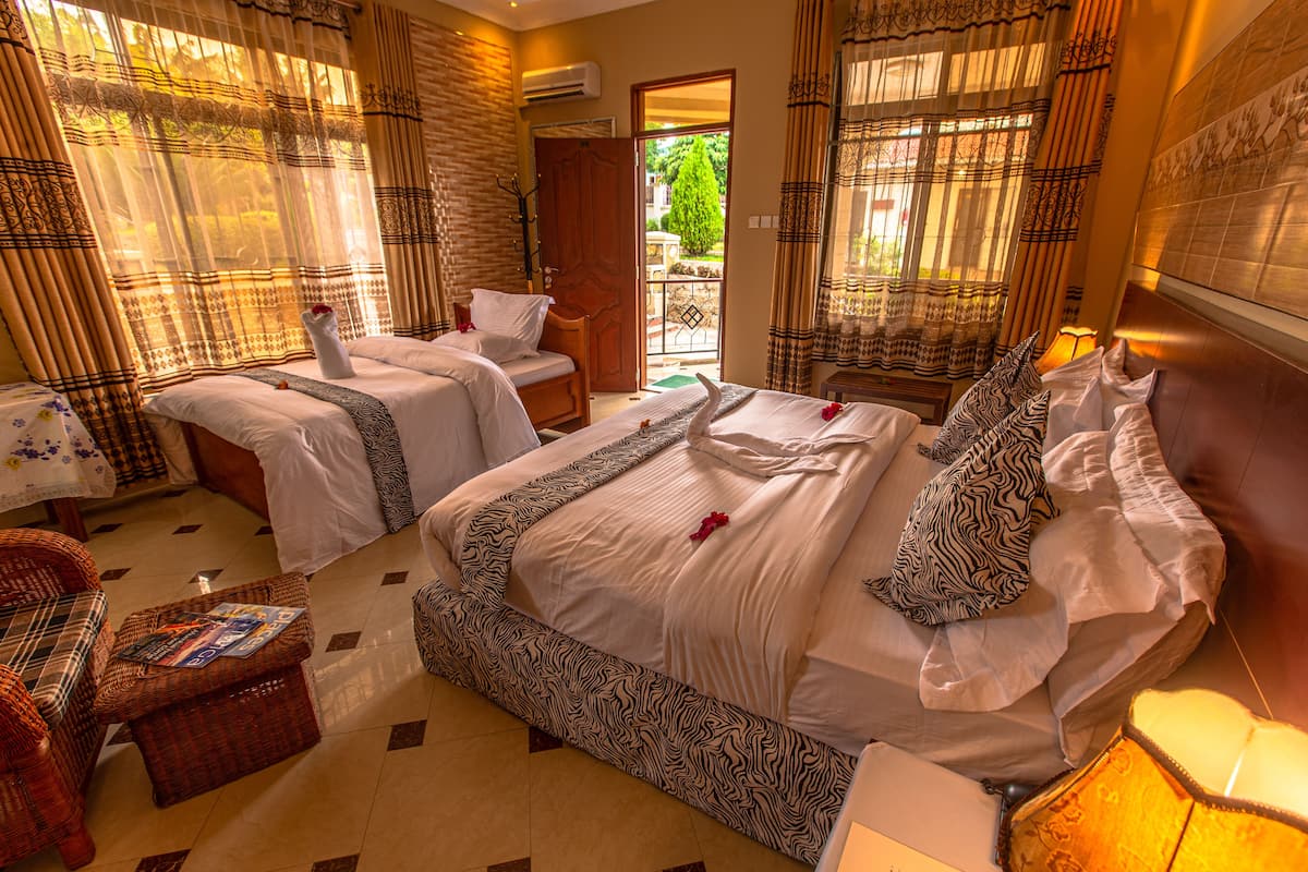 Quarto de hotel com duas camas, um espaço de estar com uma poltrona de vime e vista para o jardim pela porta.