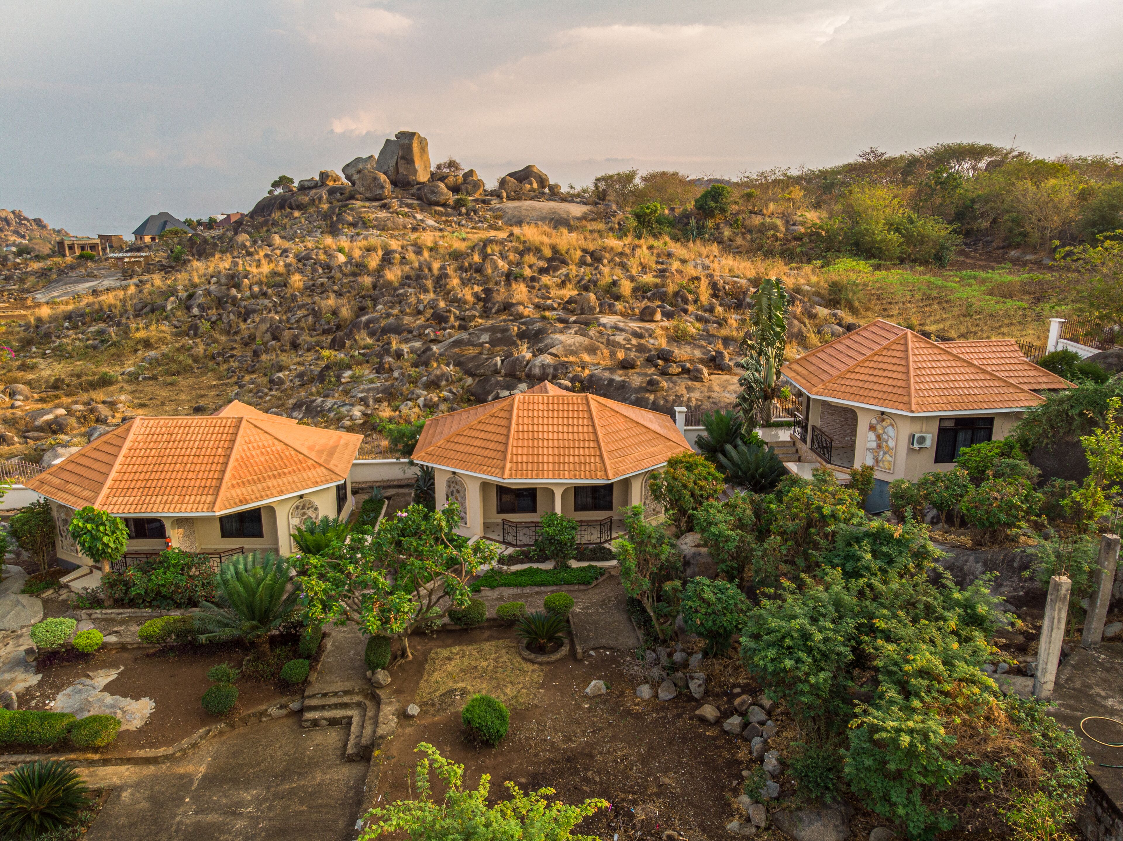 A resort with three villas featuring terracotta roofs, surrounded by landscaped gardens and a rocky hillside backdrop.