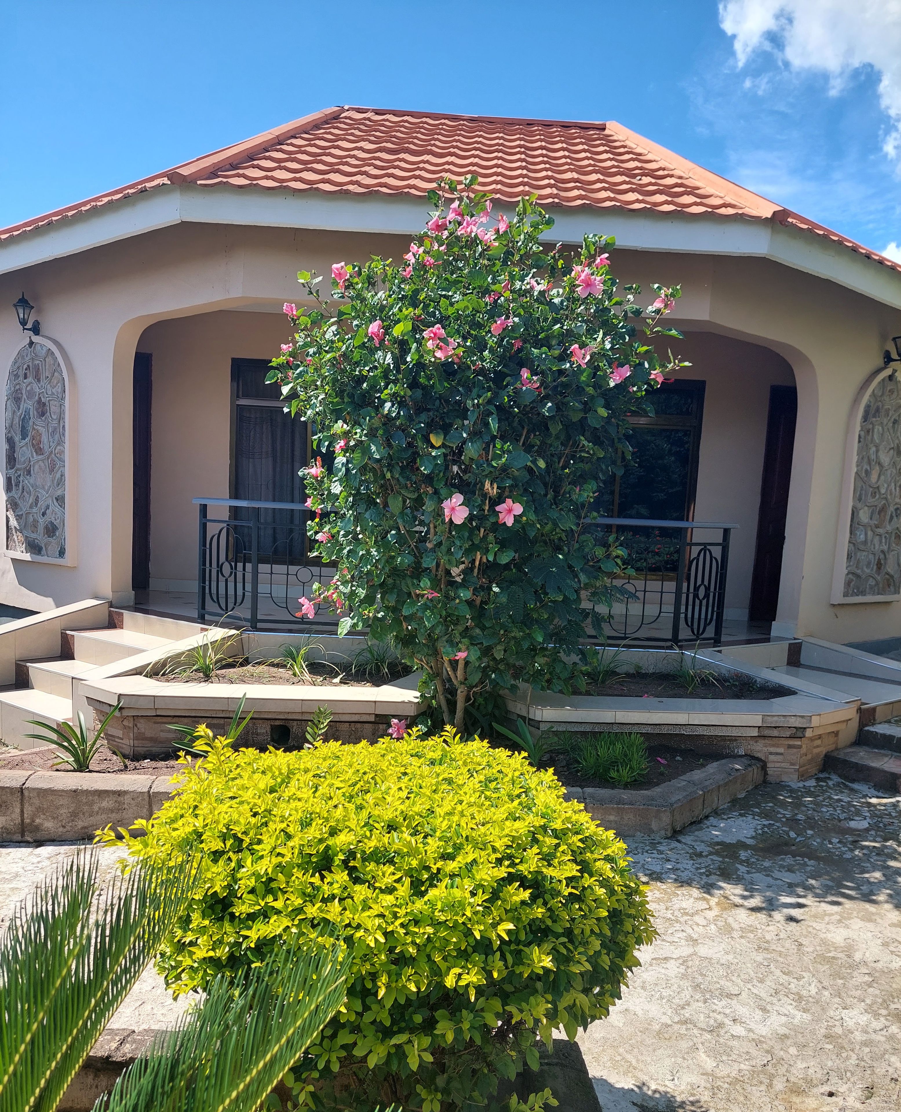 Une maison avec un toit en tuiles, un balcon et un jardin agrémenté de fleurs roses et d’arbustes verts.