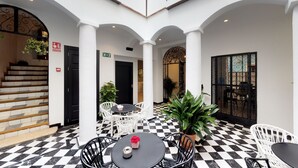 A hotel lobby with a checkered floor, white columns, and a staircase. There are tables and chairs for guests, and a large potted plant. - Madeinterranea Suites (Málaga)
