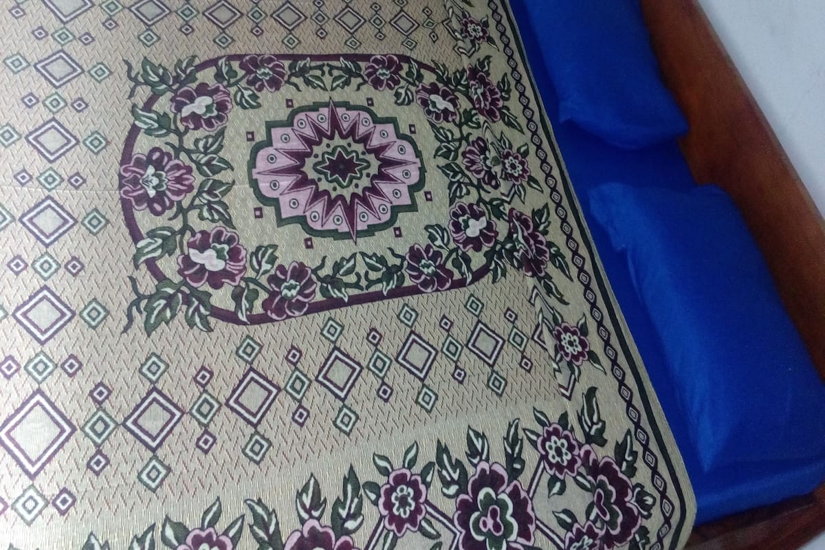 A bed with a patterned bedspread and blue sheets, a wooden headboard, and a chair in the background.