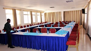 A conference room with a long table, red chairs, and a speaker on the wall. - Dlux Hotel (Nakuru)