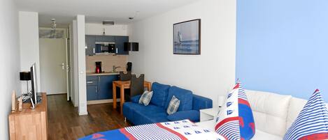 A modern hotel room with a blue and white patterned bed, a white sofa, a small dining table, and a kitchenette in the background.
