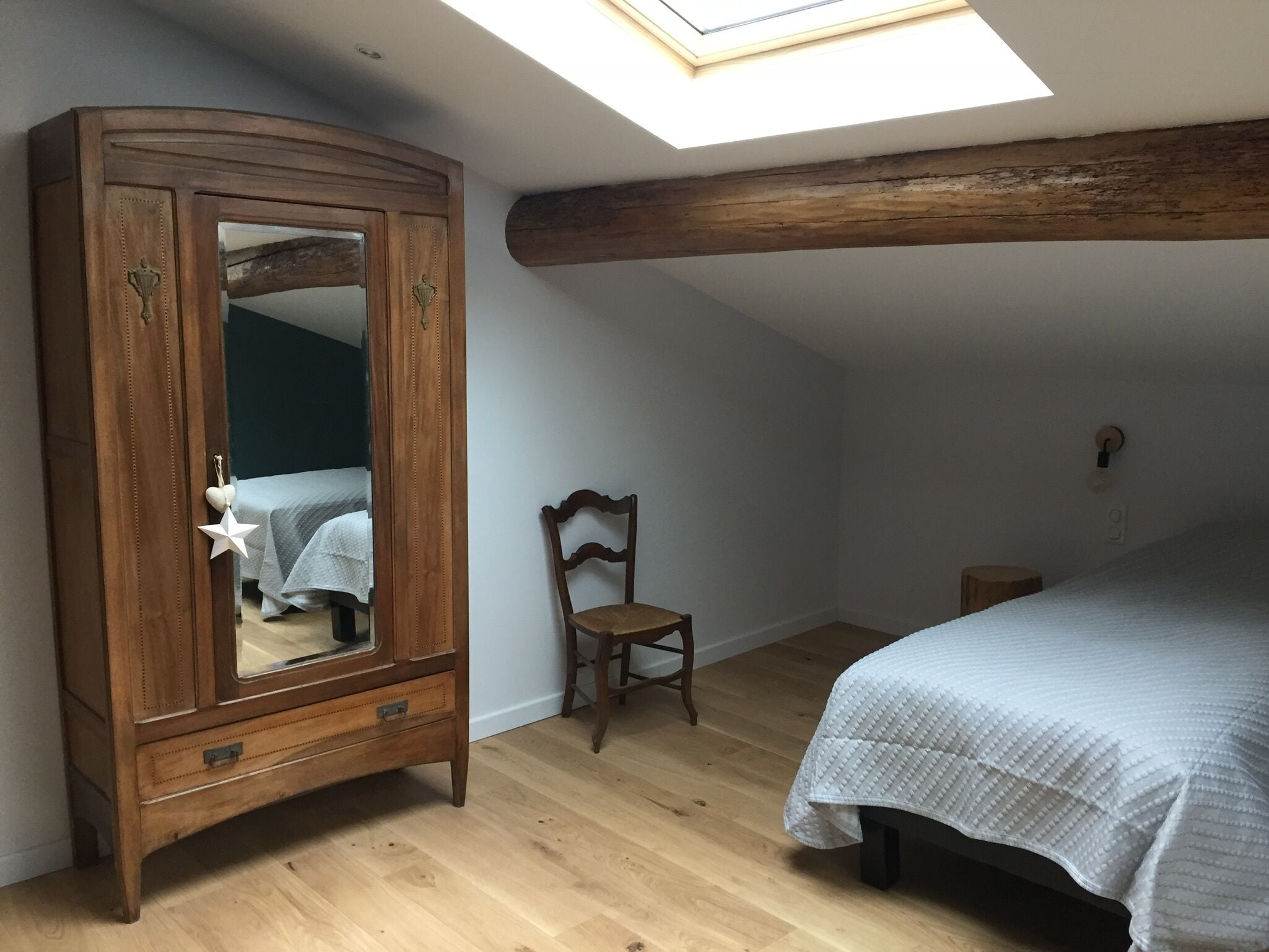 A bedroom with a skylight, wooden wardrobe, a chair, and a bed with a quilted cover.