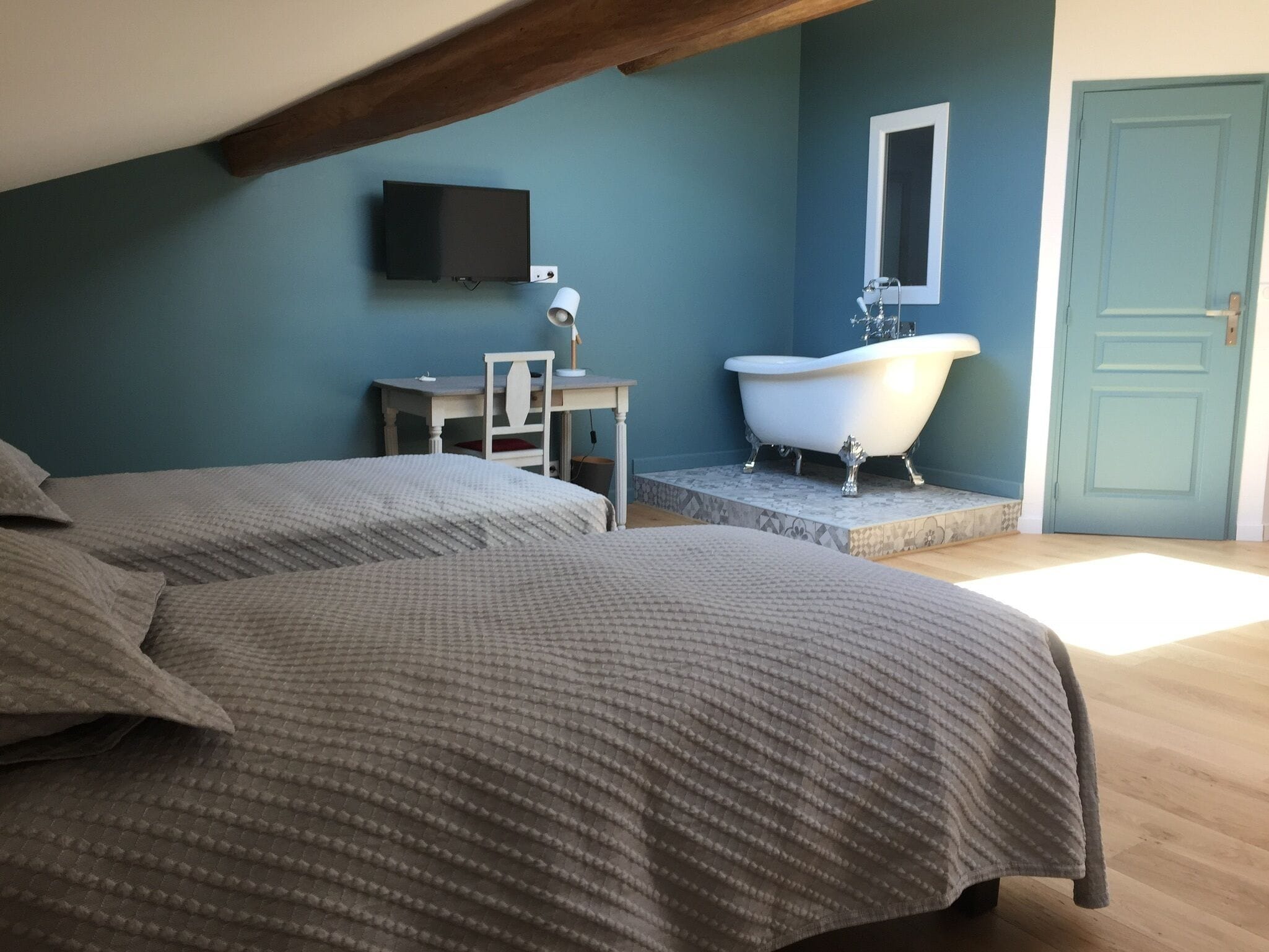 A bedroom with two beds, a desk, a flat-screen TV, a white bathtub, and a door.