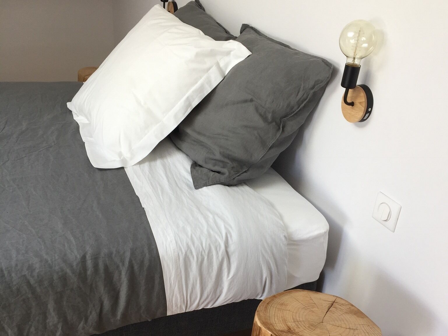 A neatly made bed with grey and white bedding, a wall-mounted light fixture, and a wooden stool.