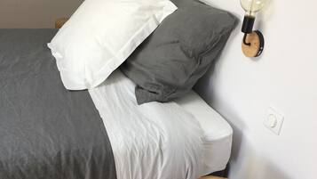 A neatly made bed with grey and white bedding, a wall-mounted light fixture, and a wooden stool.