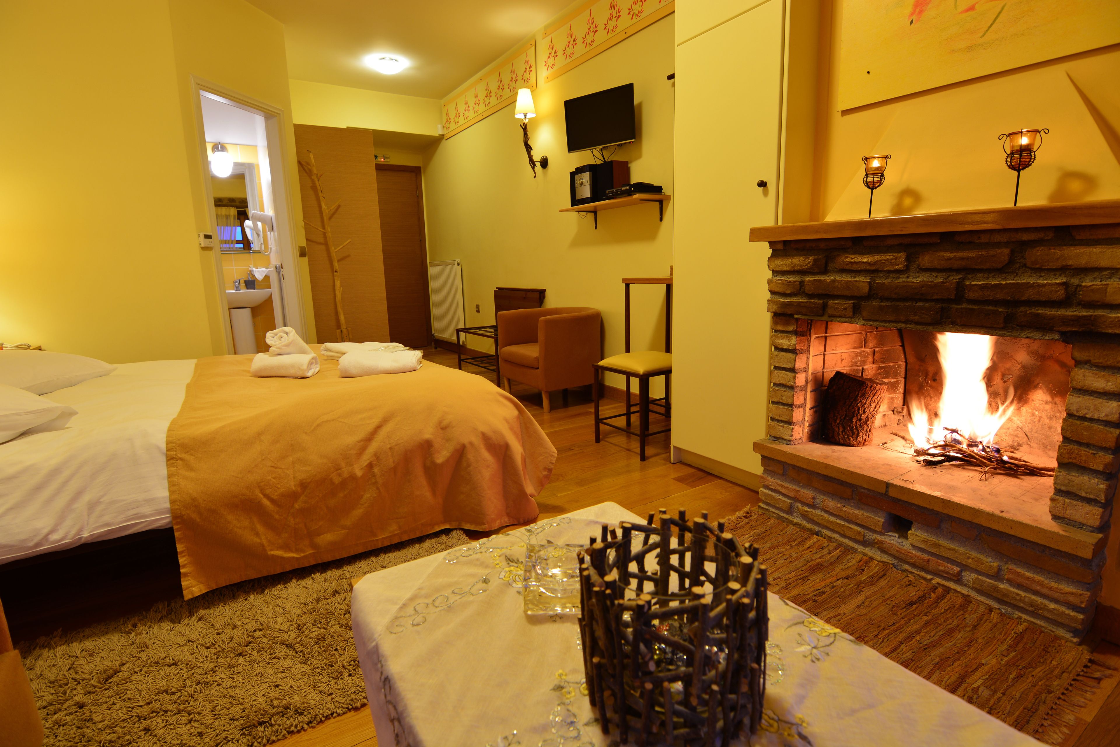 Une chambre d’hôtel comprenant un lit, un coin salon avec une cheminée, une télévision et une petite table sur laquelle est posé un vase.