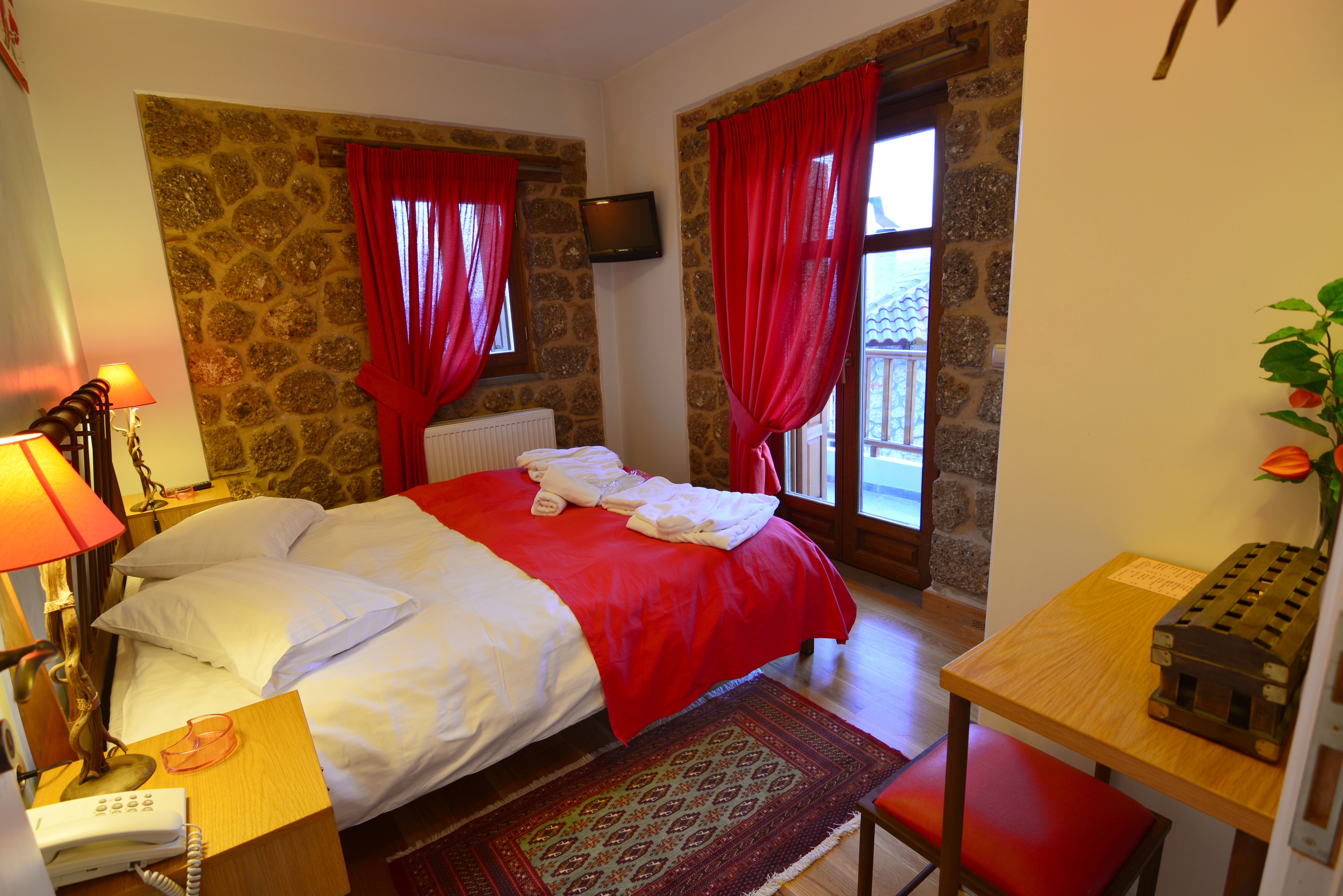 A hotel room with a bed, a desk with a telephone, a lamp, a television on the wall, and a window with red curtains.