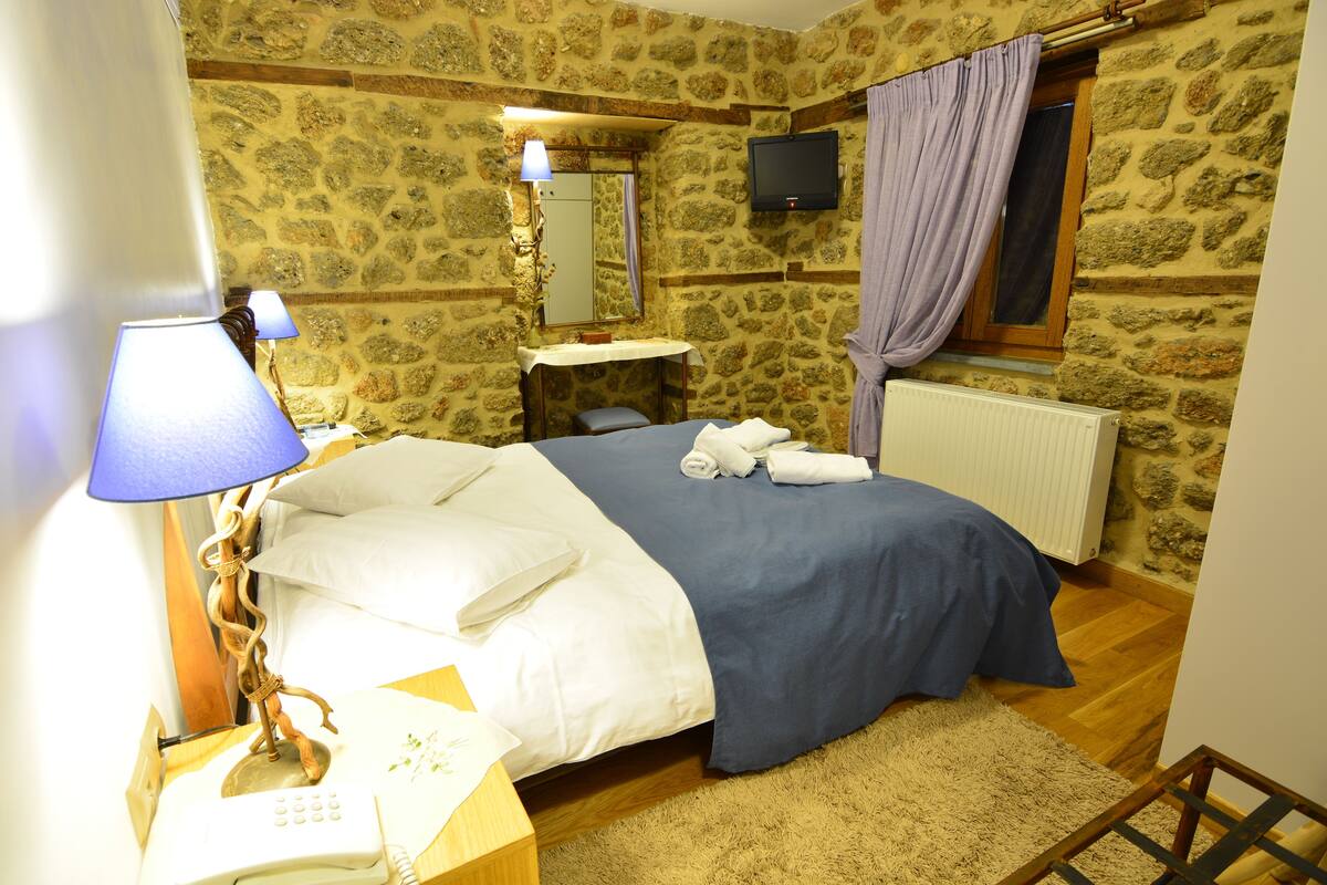 Une chambre avec un mur en pierre, un lit avec une literie blanche et bleue, une lampe de chevet, un téléphone, un miroir et un téléviseur fixé au mur.