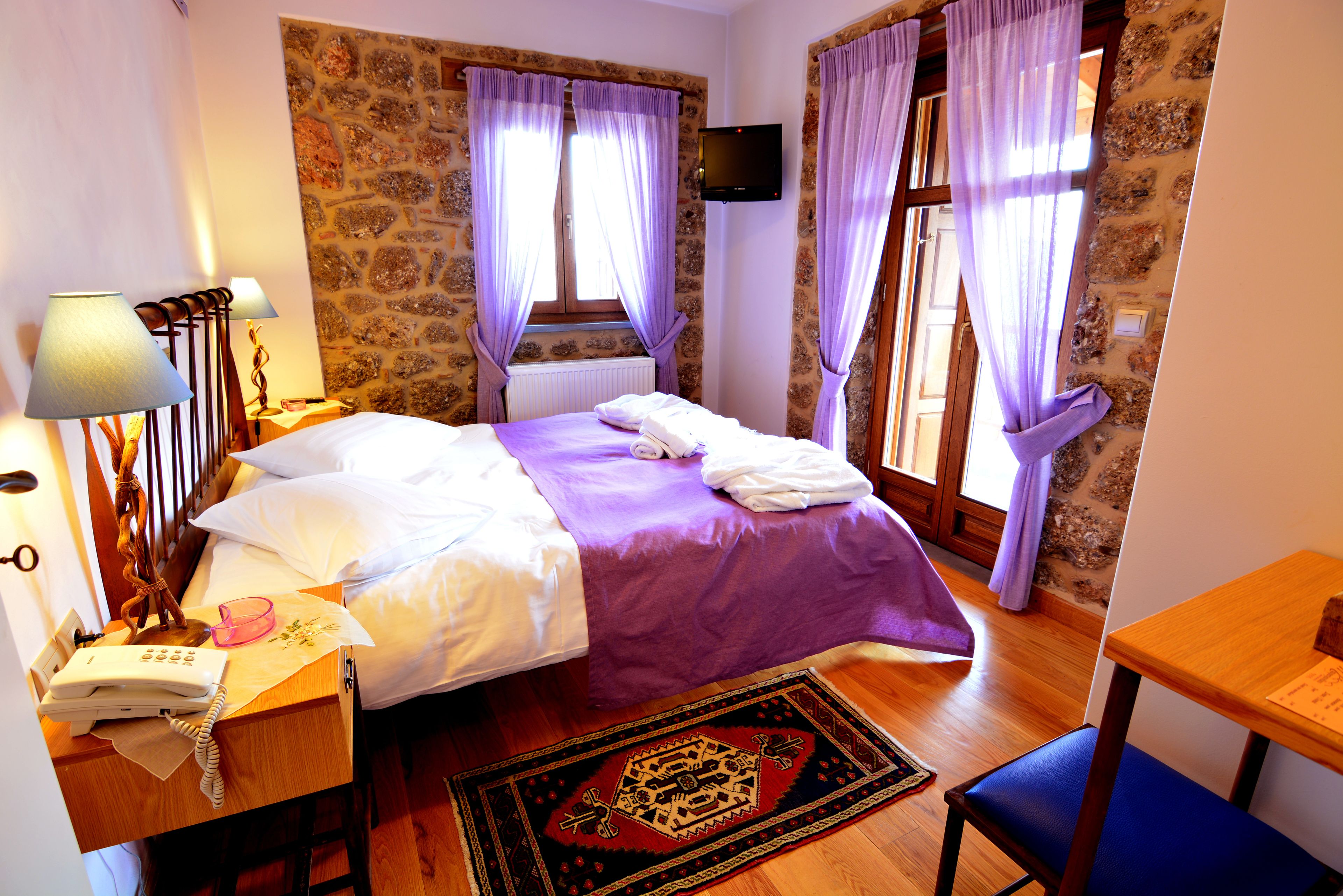 Un dormitorio con cama, escritorio con teléfono, silla, televisor en la pared y ventana con cortinas.