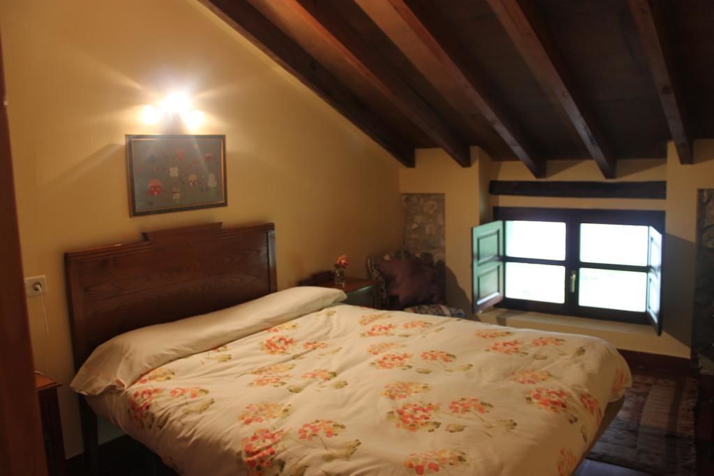 Un dormitorio con cabecera de madera, colcha estampada con motivos florales, mesita de noche, una silla y una ventana con luz diurna.