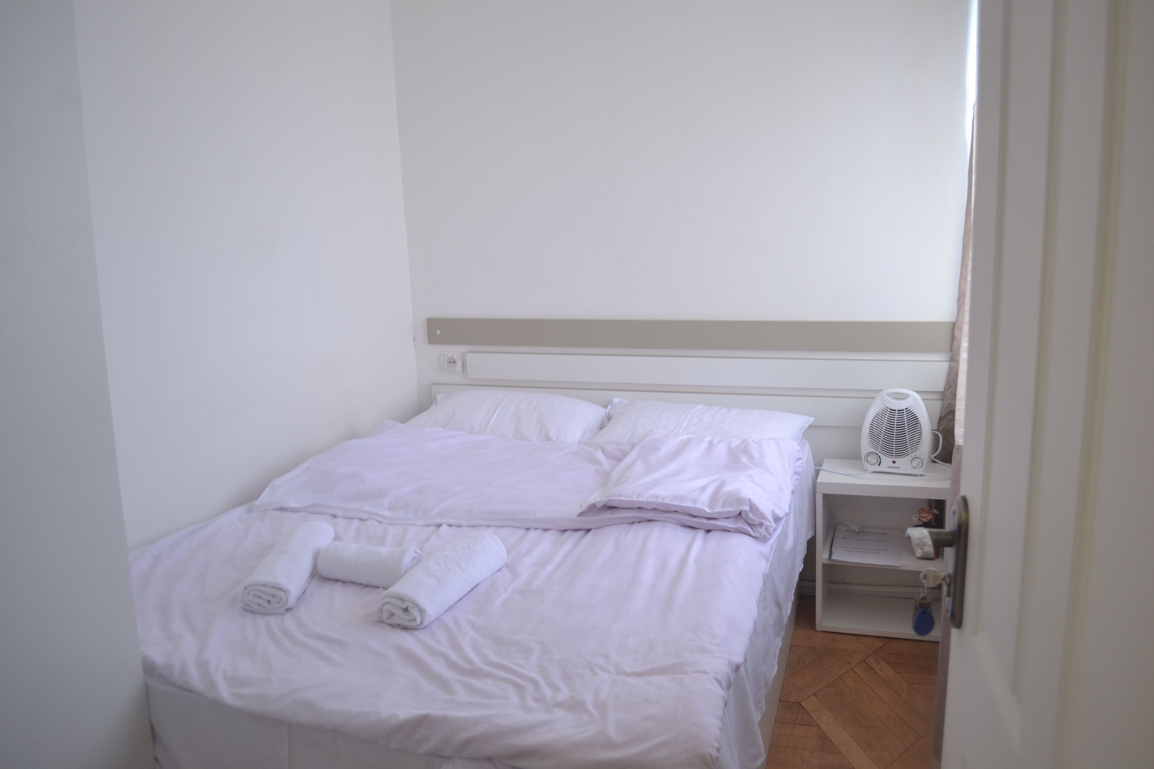 A neatly made bed with white linens and rolled towels, a bedside table with a fan, and a small shelf with a book and a keychain.