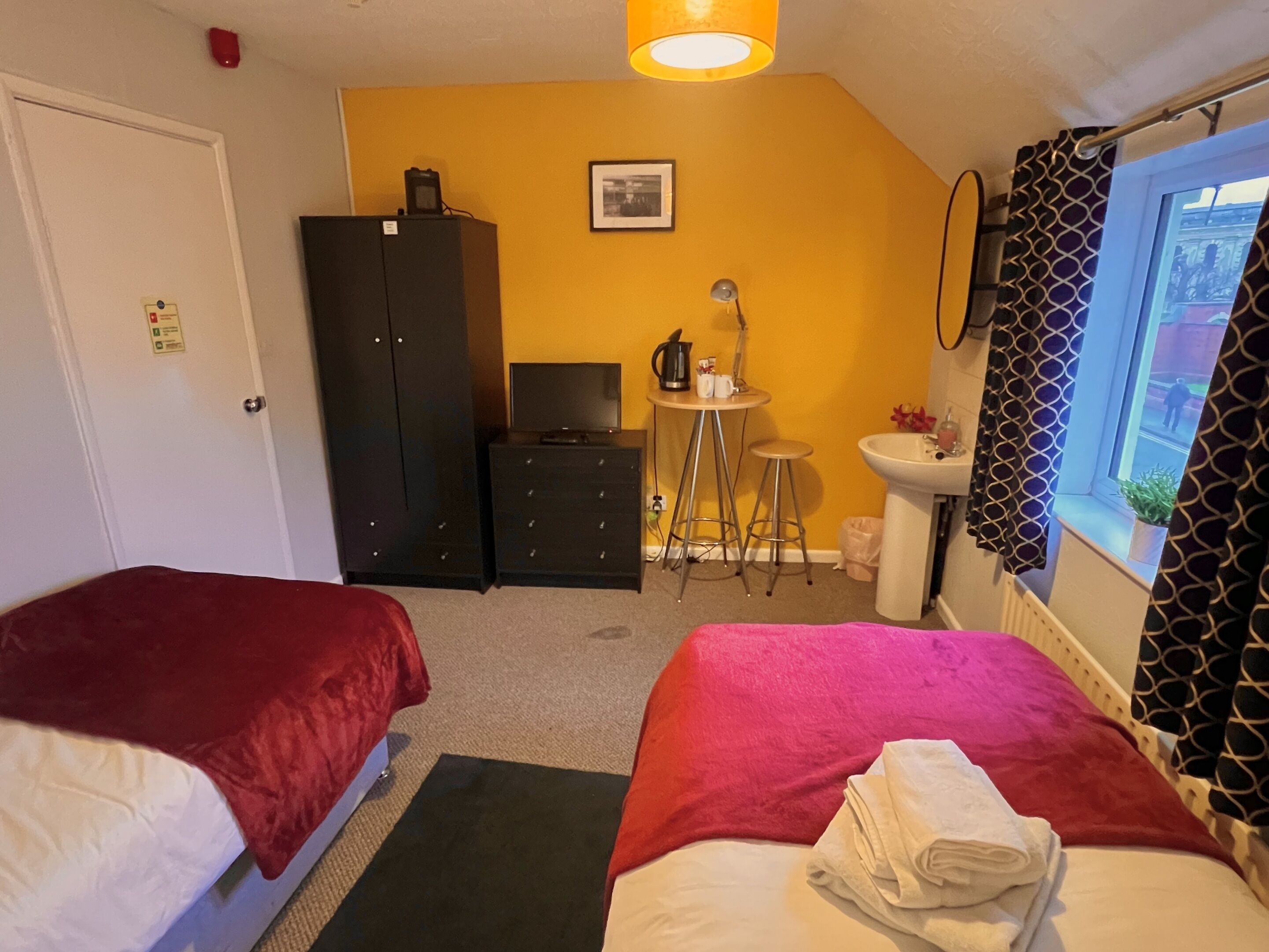 Ein Zimmer mit zwei Betten, einem Schreibtisch, einem Stuhl, einem kleinen Tisch mit Wasserkocher, einem Waschbecken und einem Fenster mit Vorhängen.