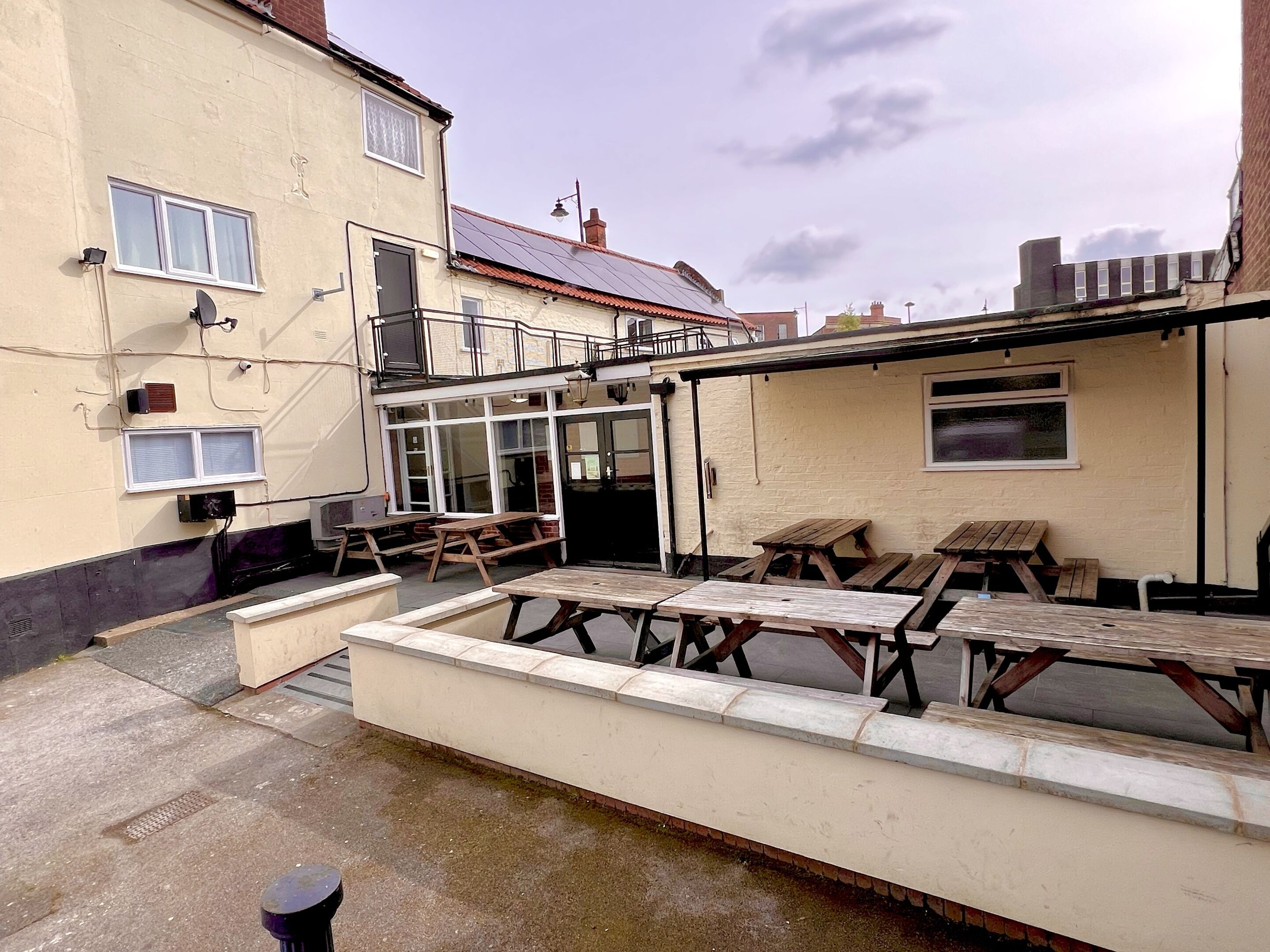 A patio area with wooden tables and benches, a small trash bin, and a building with a tiled roof and windows.