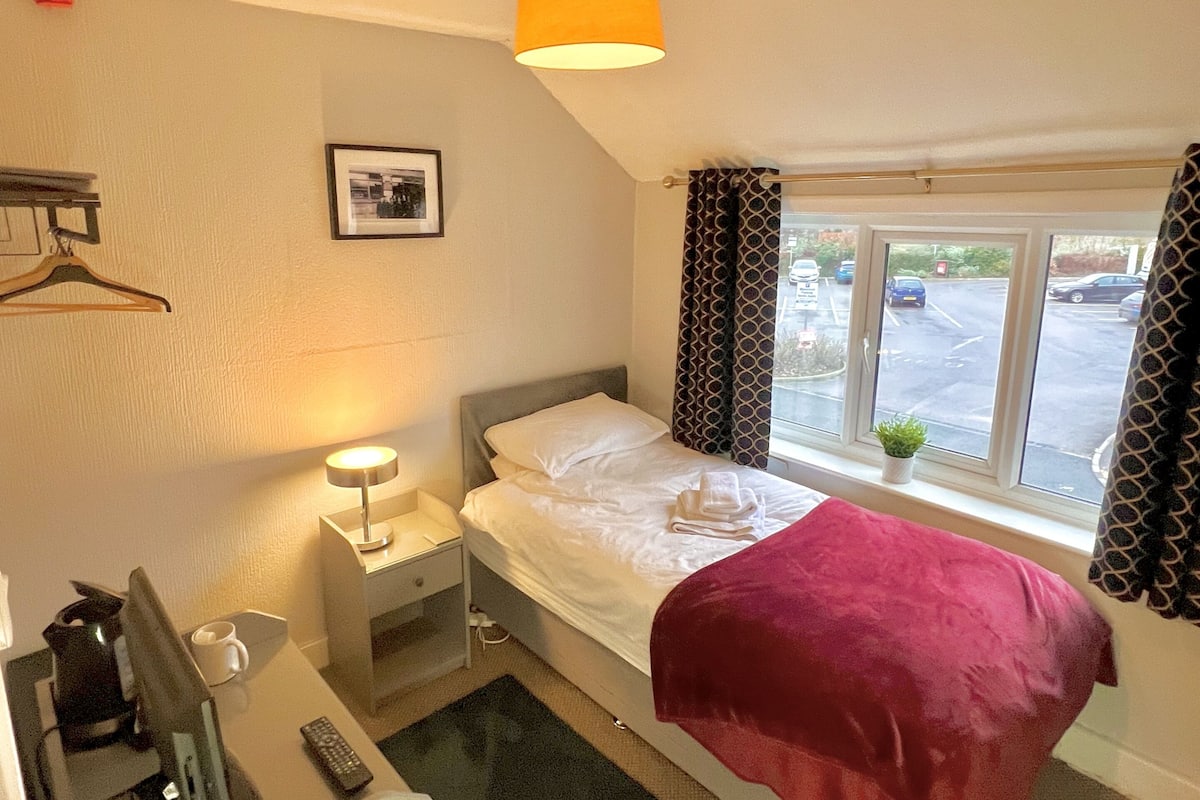 Une chambre d’hôtel avec un lit, une table de chevet avec une lampe, un porte-manteau mural, une fenêtre avec des rideaux et une vue sur le parking extérieur.
