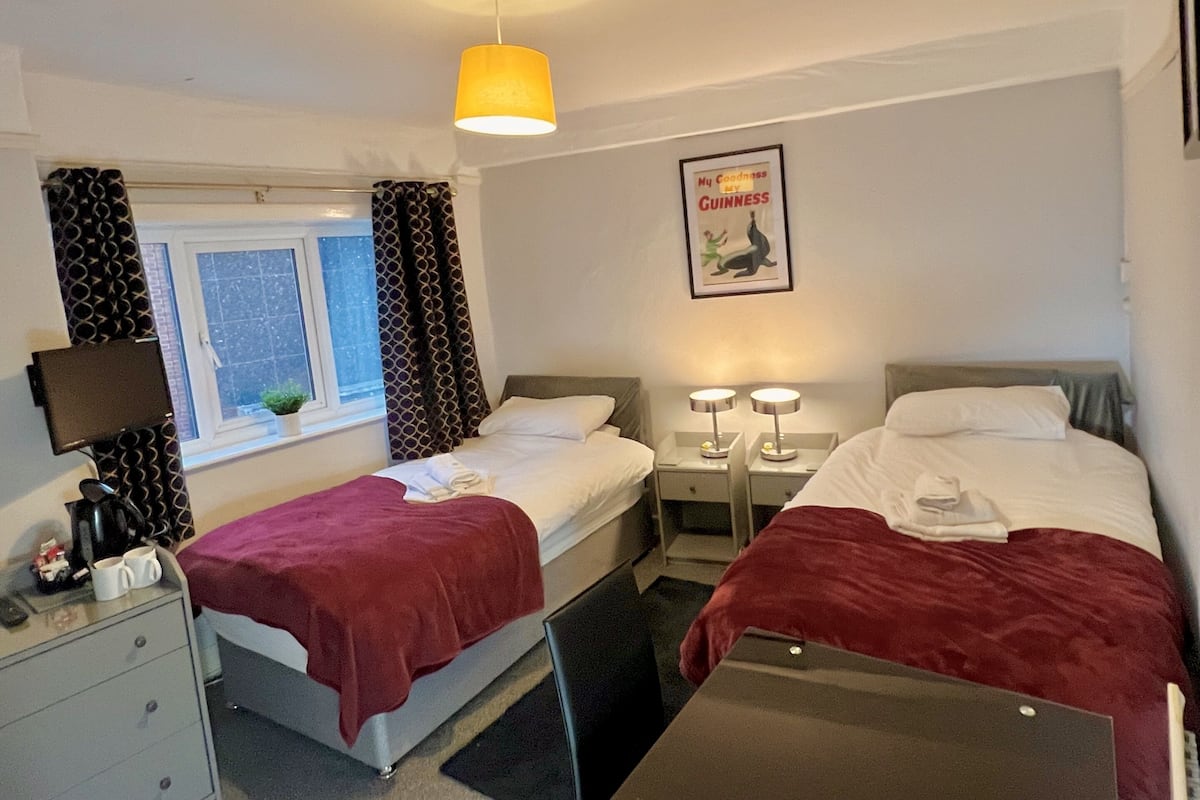 Une chambre d’hôtel avec deux lits, une fenêtre avec des rideaux, une affiche encadrée au mur et un bureau avec une lampe.