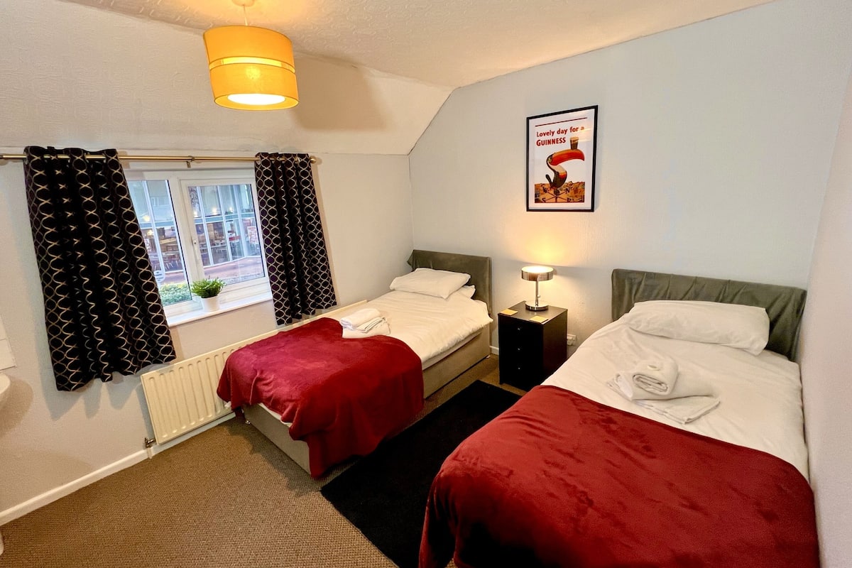 Une chambre d’hôtel avec deux lits, une fenêtre avec des rideaux à motifs, un radiateur et une affiche encadrée au mur.