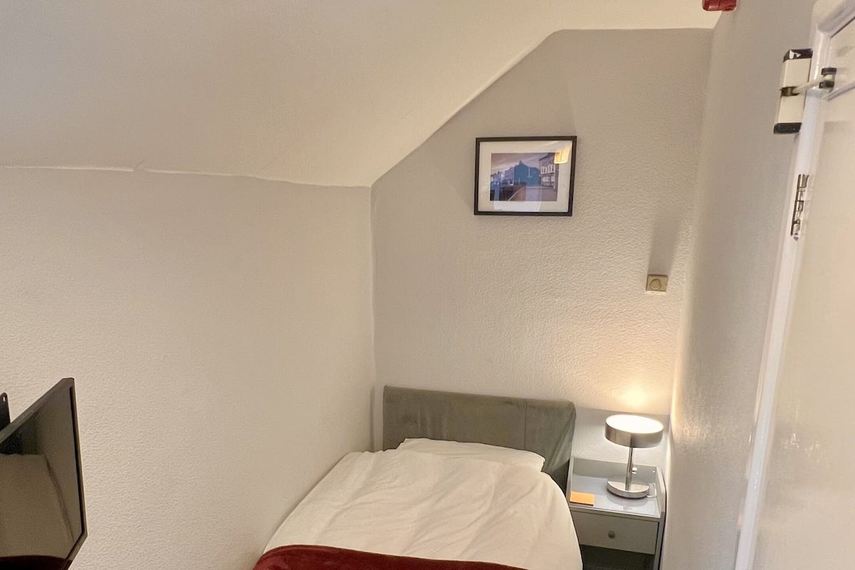 Une petite chambre d’hôtel, propre et bien rangée, avec un lit simple, une table de chevet avec une lampe, un tableau encadré au mur et un plafond blanc.