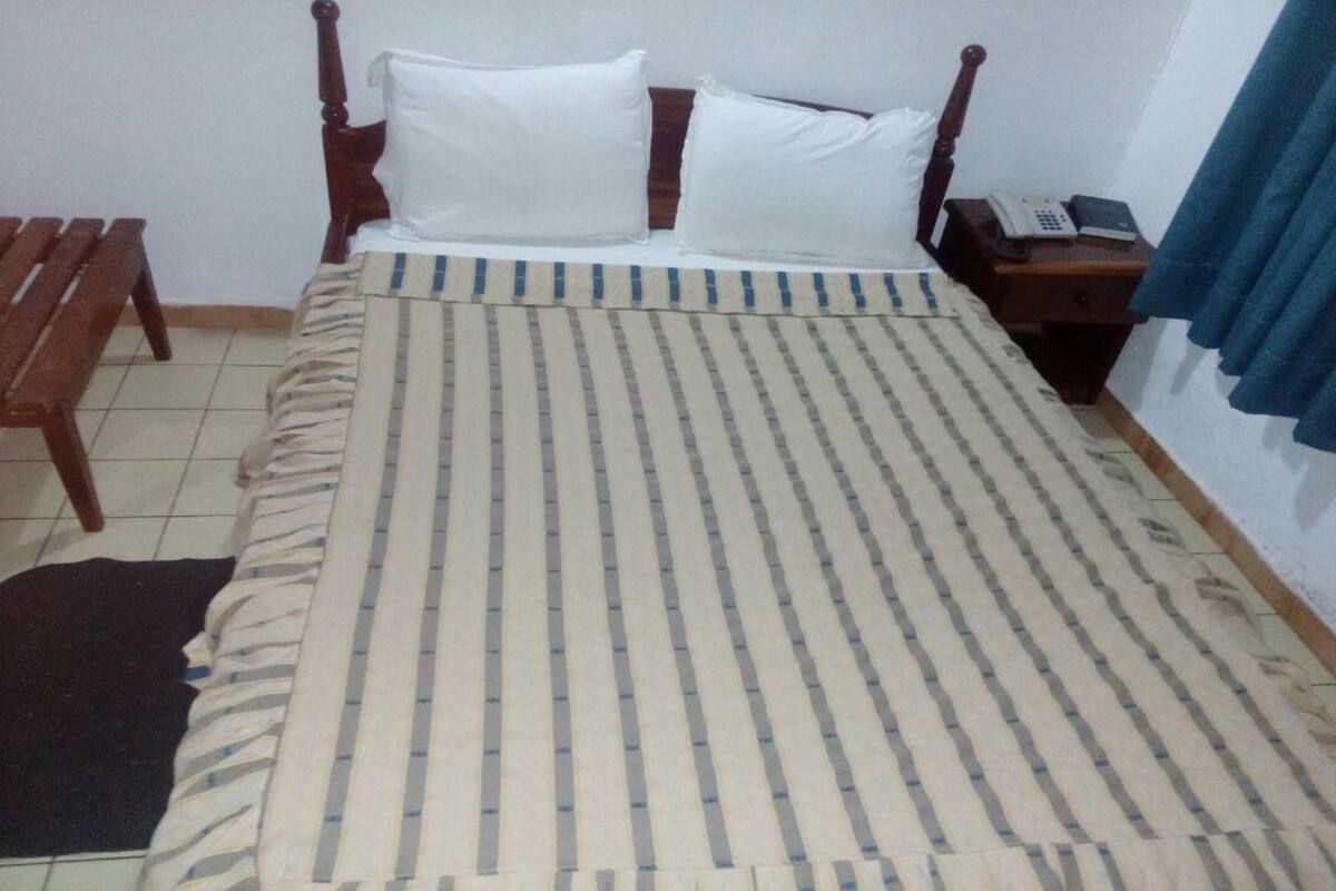 A single bed with a striped bedspread, a wooden nightstand with a telephone, and a bench to the left.