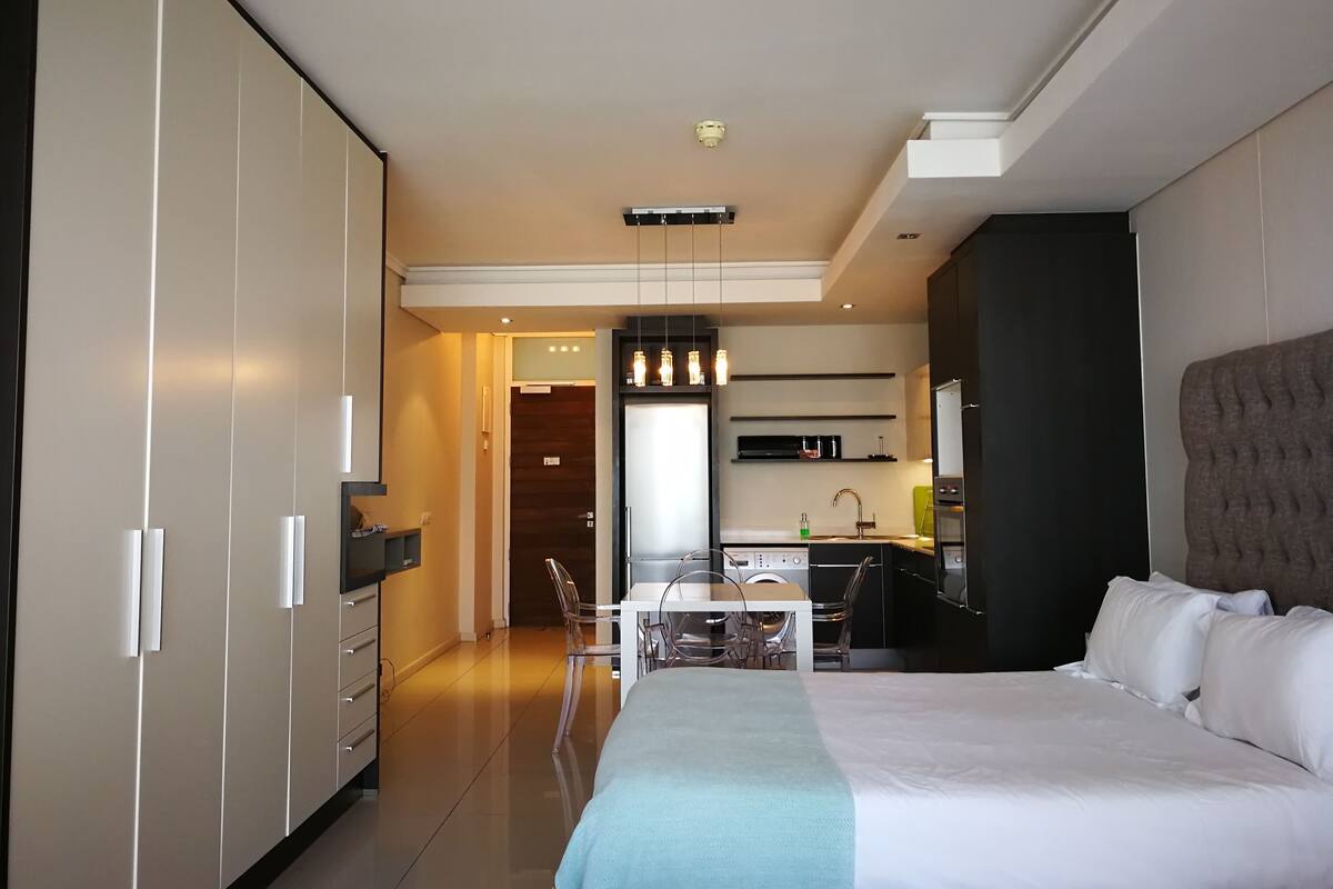 A modern hotel room with a large bed, a desk, and a kitchenette.