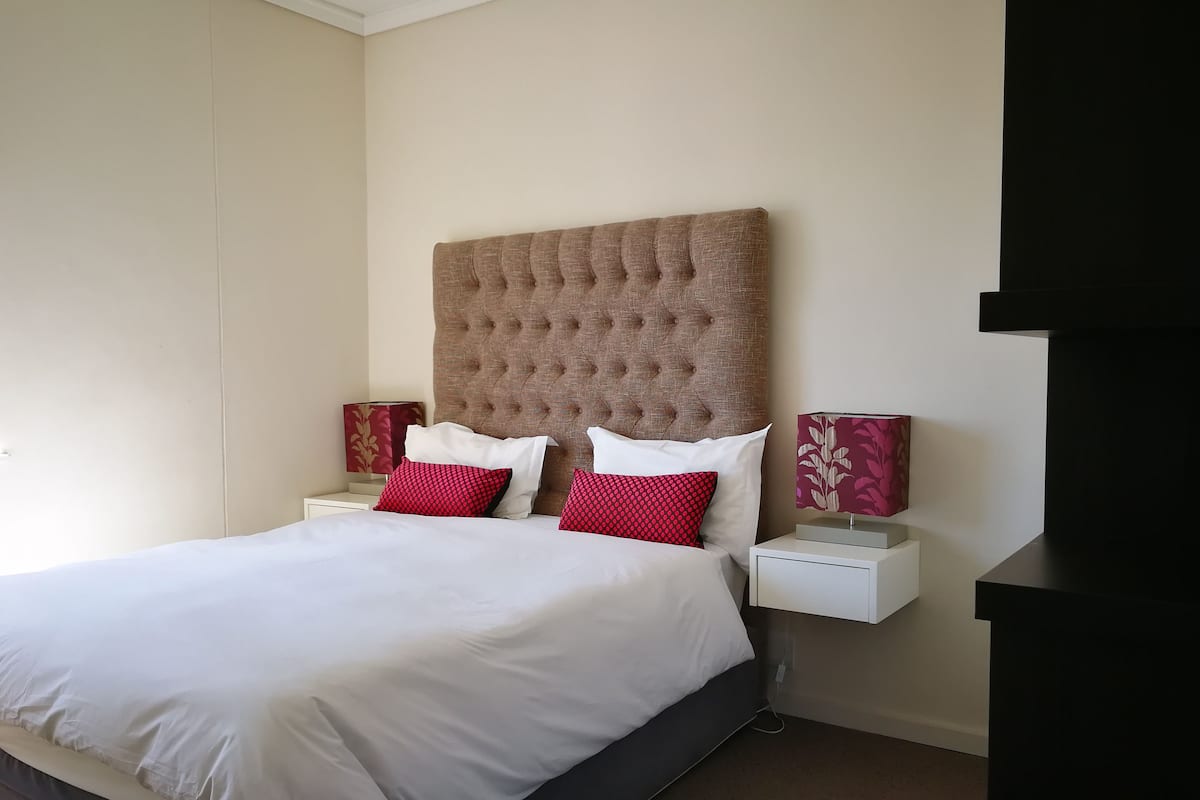 A neatly made bed with white and red pillows, a tufted headboard, and two bedside tables with lamps and decorative items.