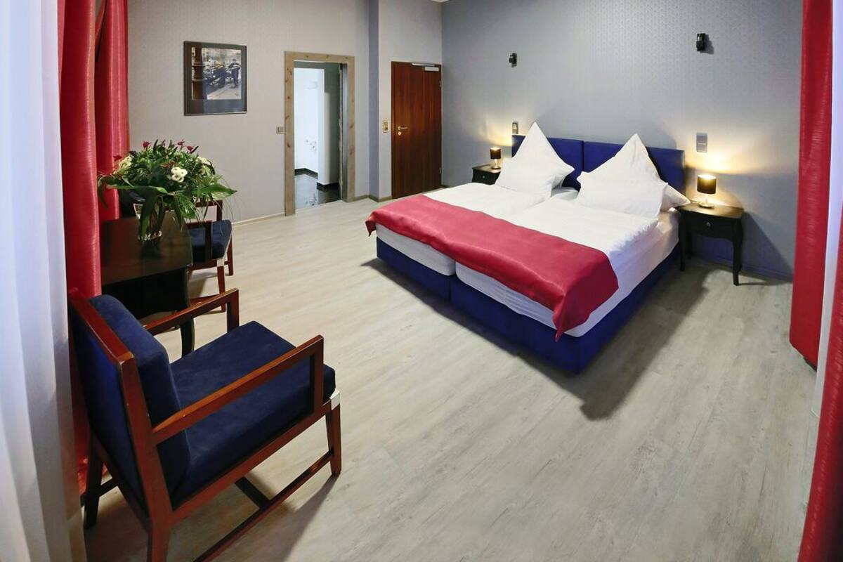 A hotel room with a bed, bedside tables, a chair, a small table with a vase of flowers, and a door leading to another room.