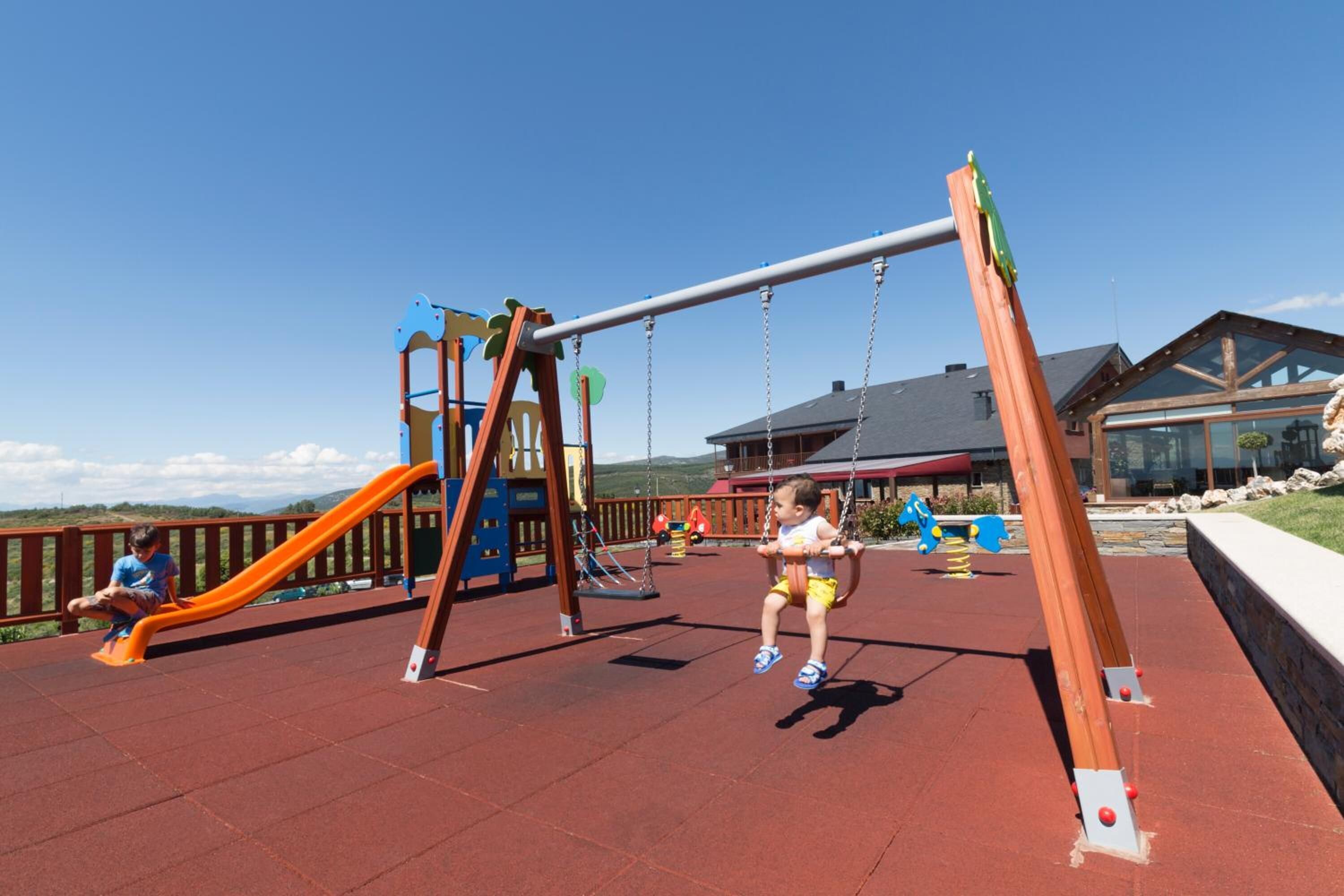 Un parque infantil con columpios y toboganes, una estructura de madera, y un edificio al fondo.